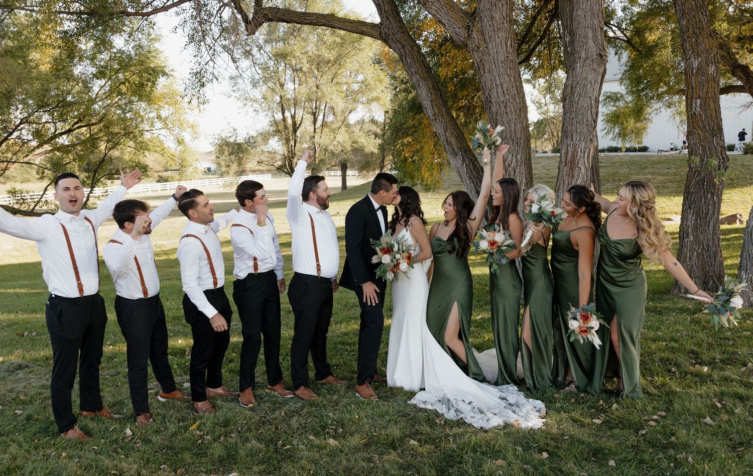 Wedding party in green dresses and suspenders poses joyfully under trees.