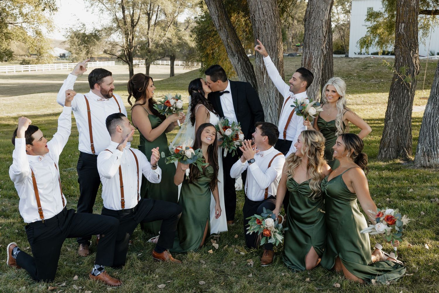 A wedding party in green dresses and suspenders poses playfully under trees outdoors.