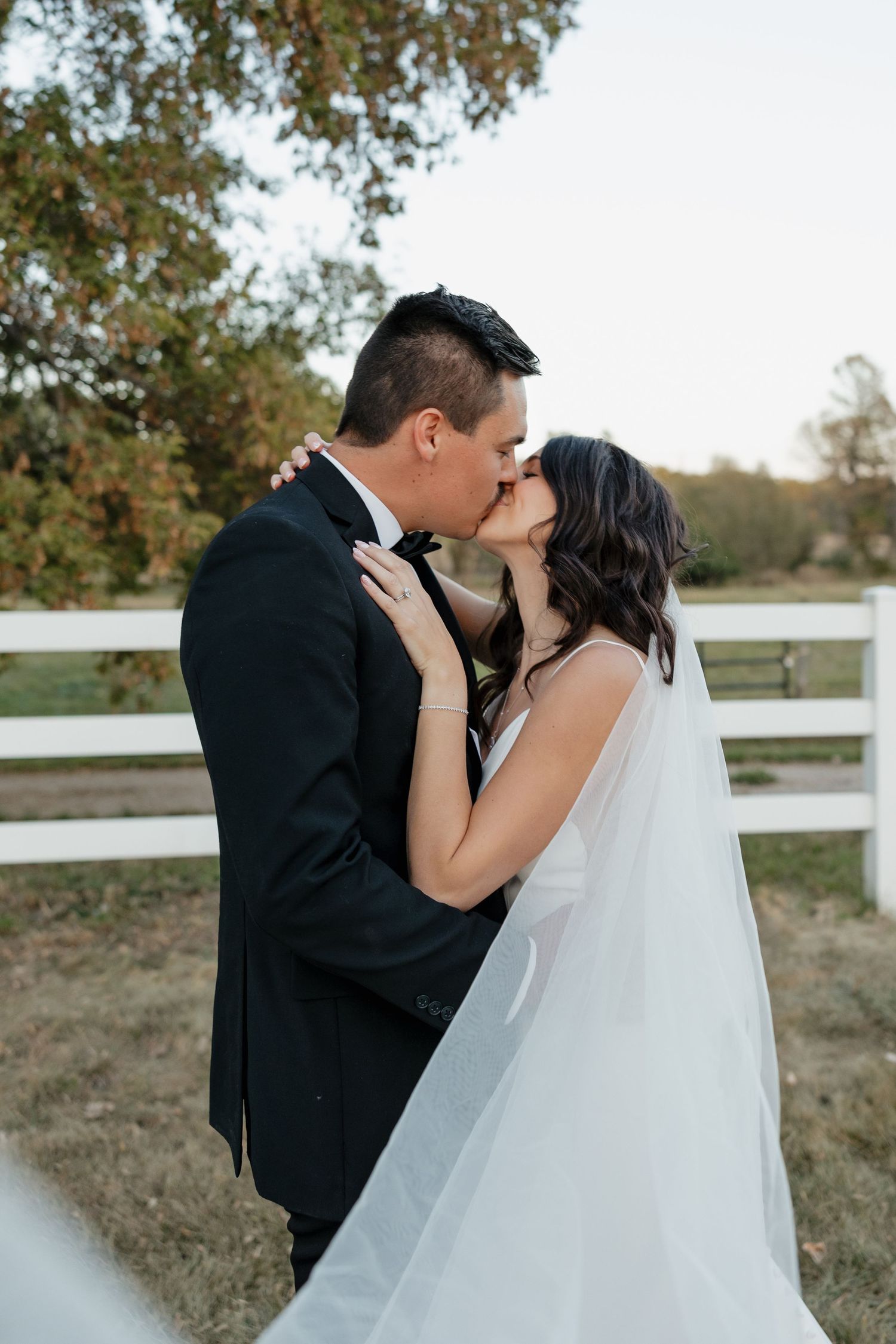 Intimate wedding kiss portrait by white fence in natural outdoor setting.