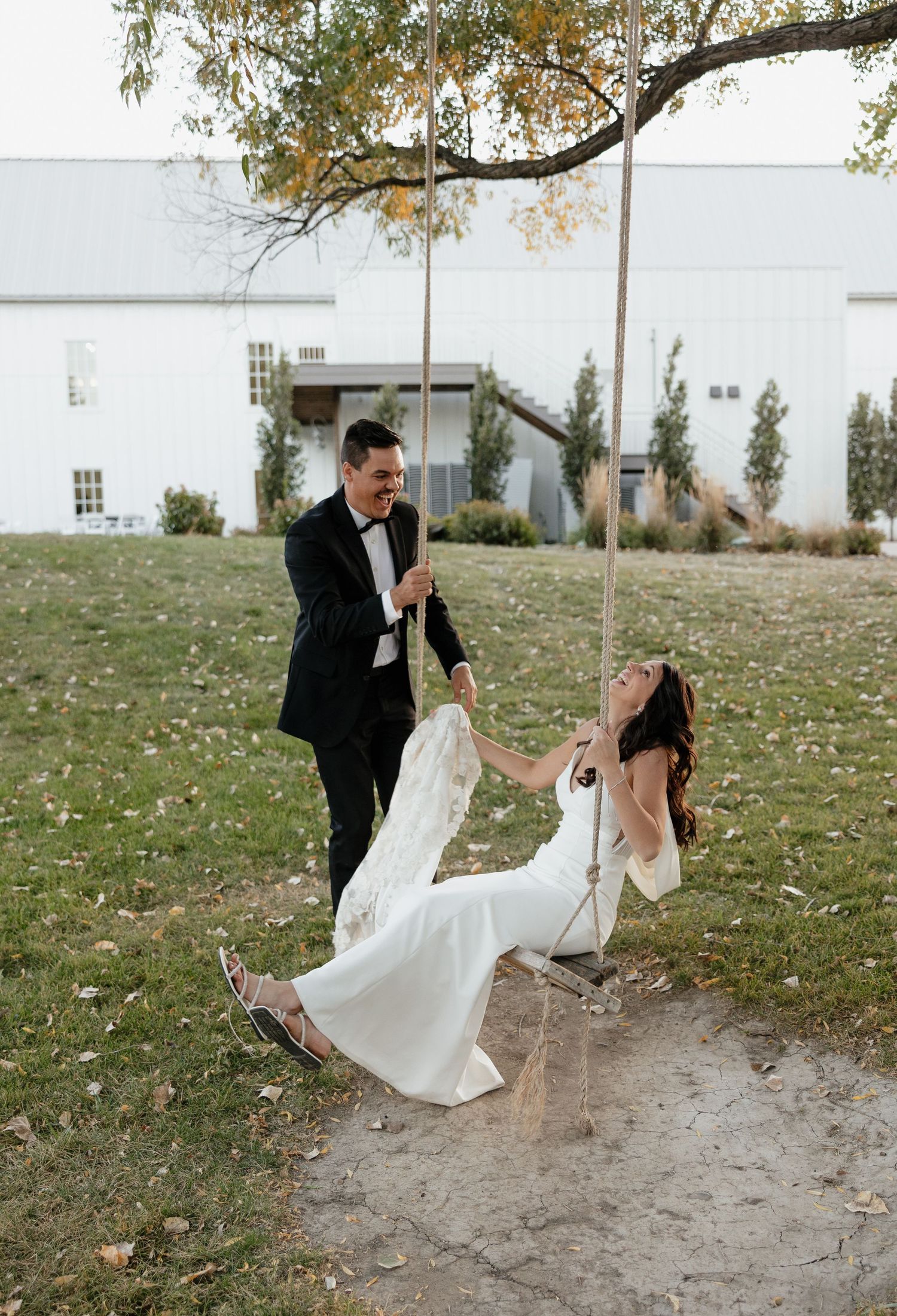 A couple shares a playful moment on a tree swing during their wedding photoshoot on a grassy lawn.