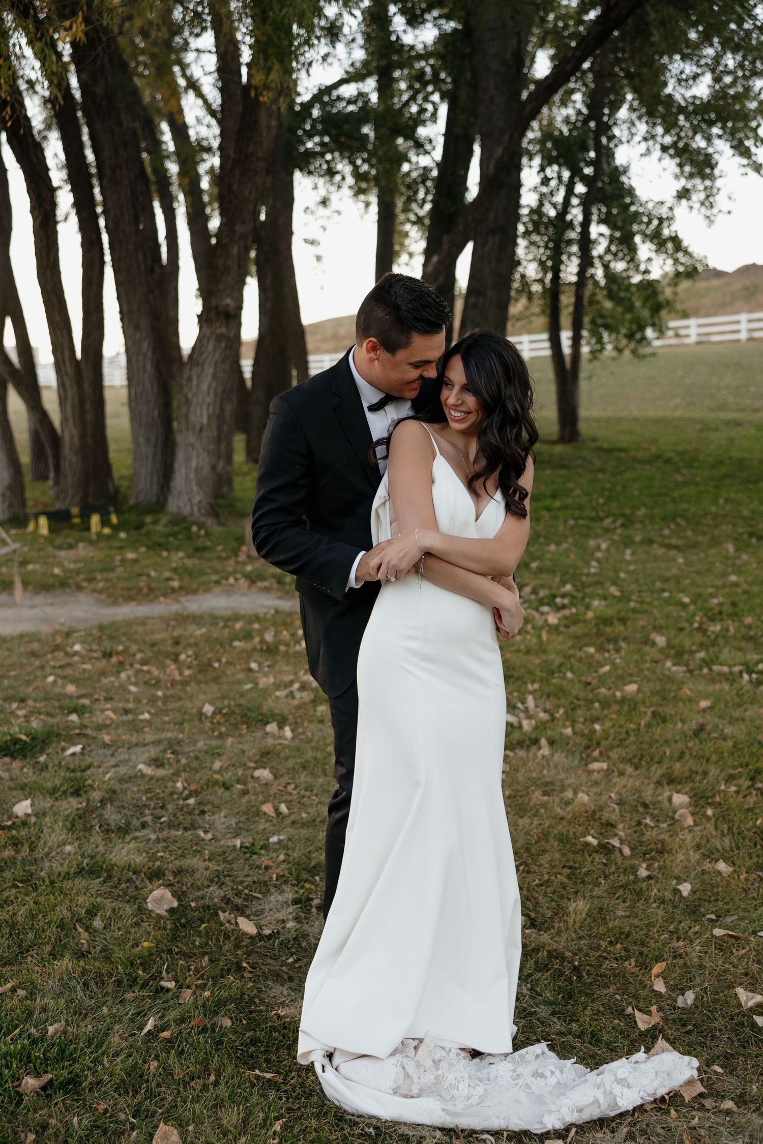 A romantic wedding portrait of a couple embracing outdoors with trees and white fencing in the background.