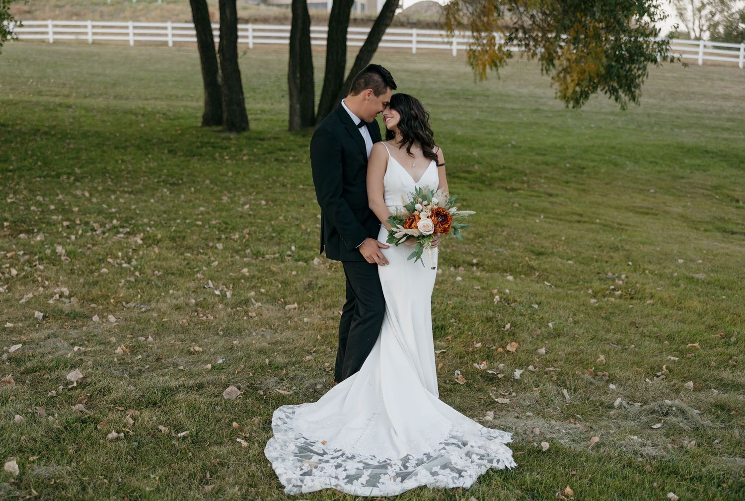 A wedding couple poses beneath autumn trees with a white fence visible in the background.