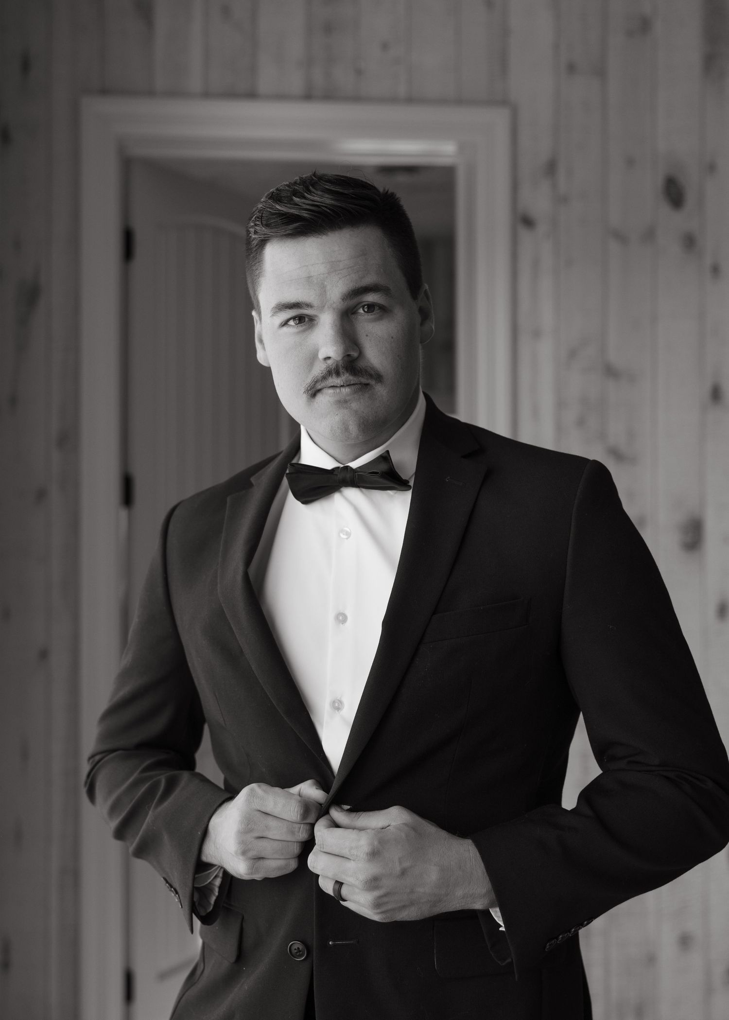 Person wearing a classic black tuxedo with bowtie stands in front of wooden paneled wall.