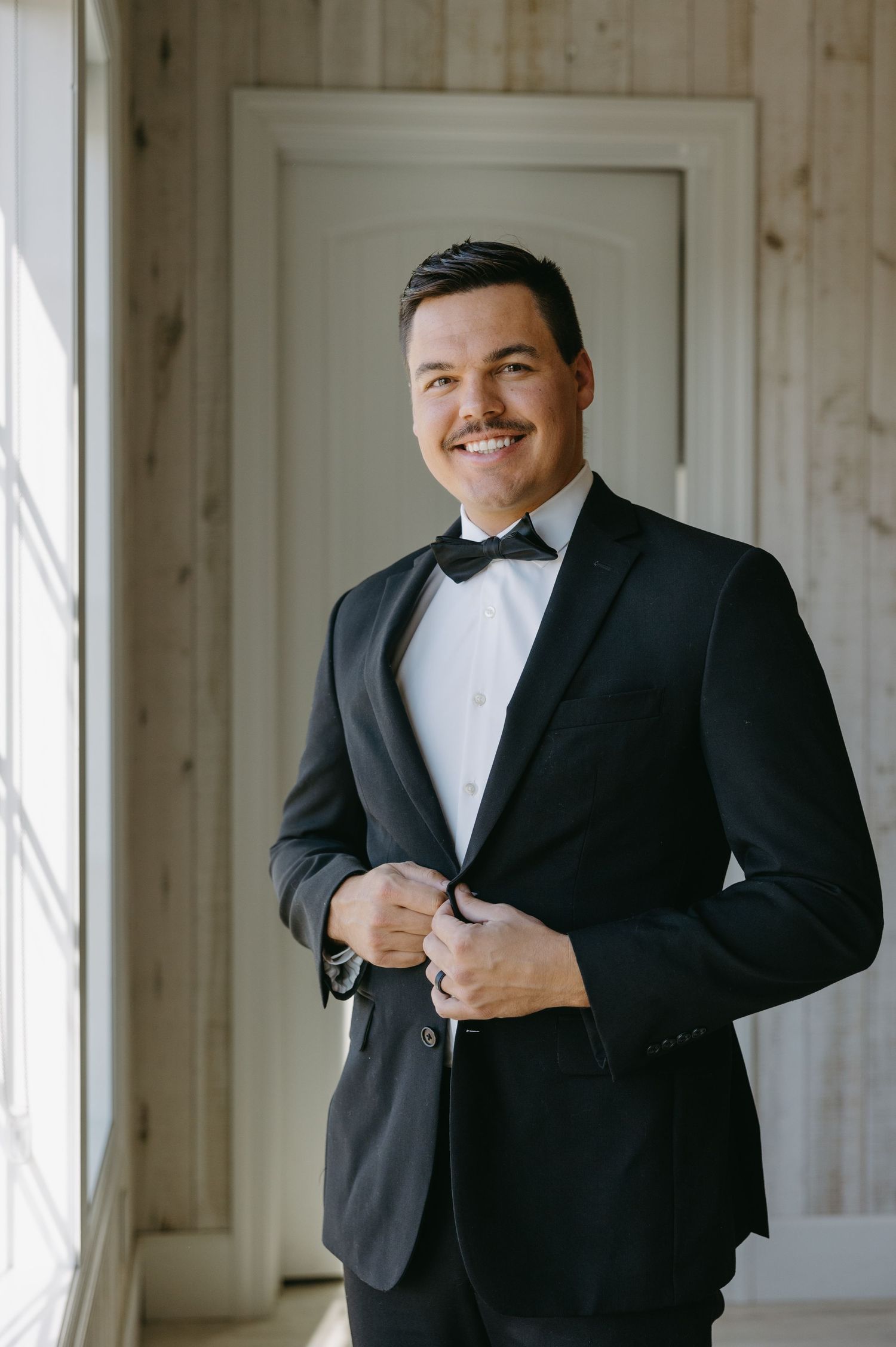 Formal portrait of someone in a black tuxedo and bowtie against wooden paneled wall.