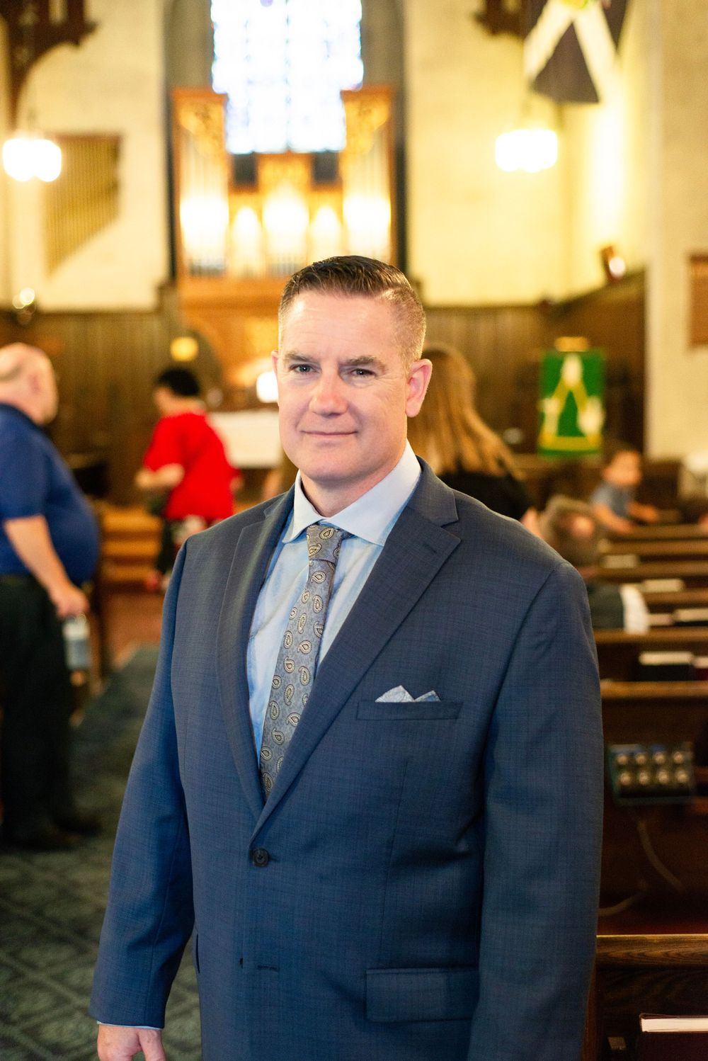 A guest wearing a navy suit and gray tie stands in a church sanctuary with wooden pews and warm lighting.