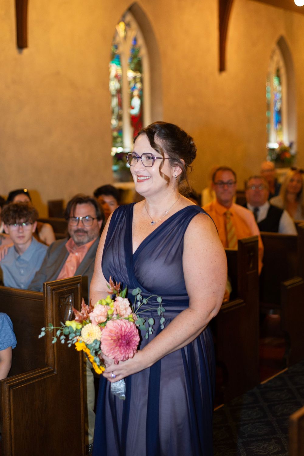 A bridesmaid in a navy blue dress holding a pink and yellow bouquet stands smiling in a church with stained glass windows.