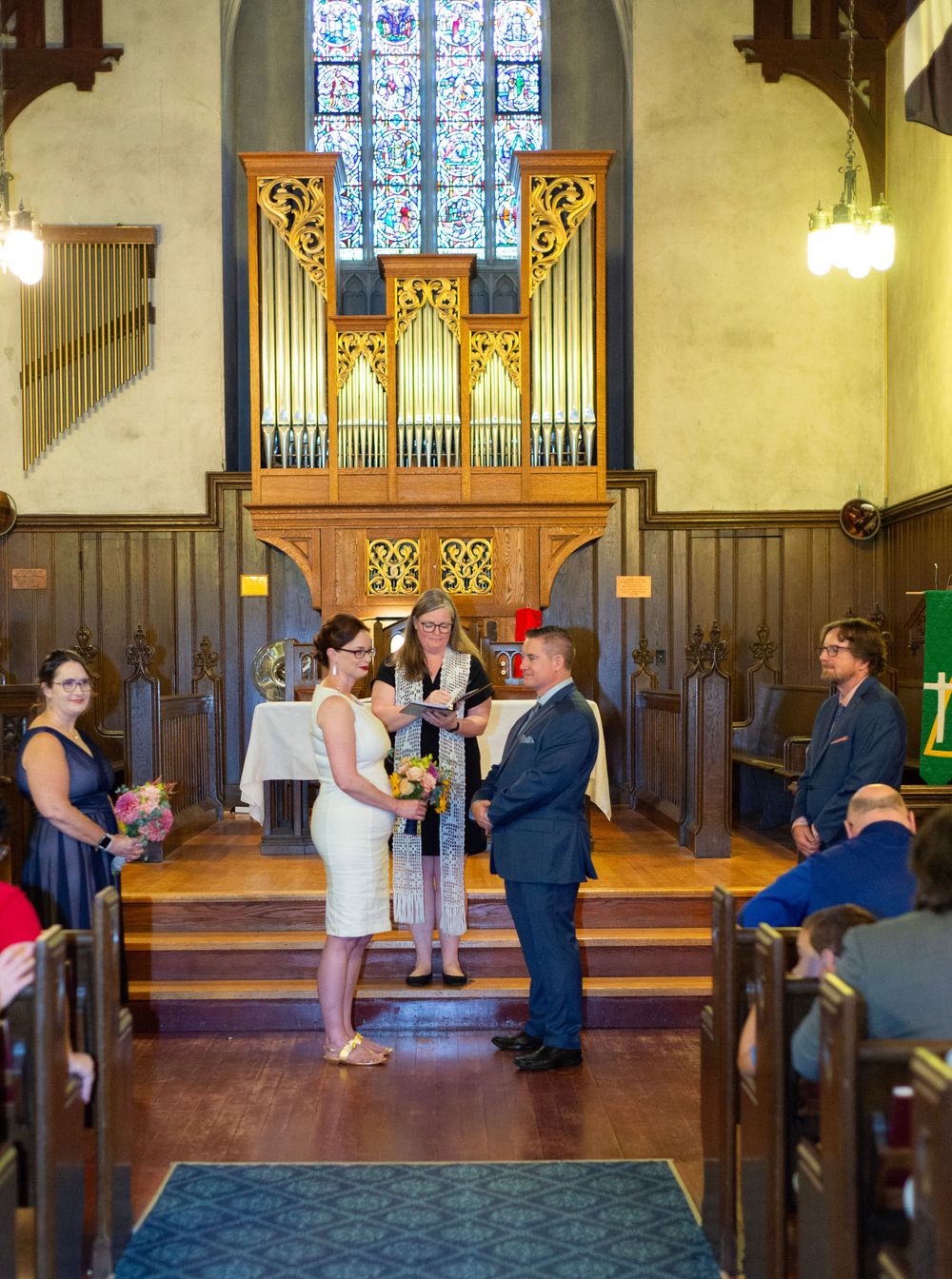 A wedding ceremony taking place in a chapel with a beautiful pipe organ and stained glass windows in the background.