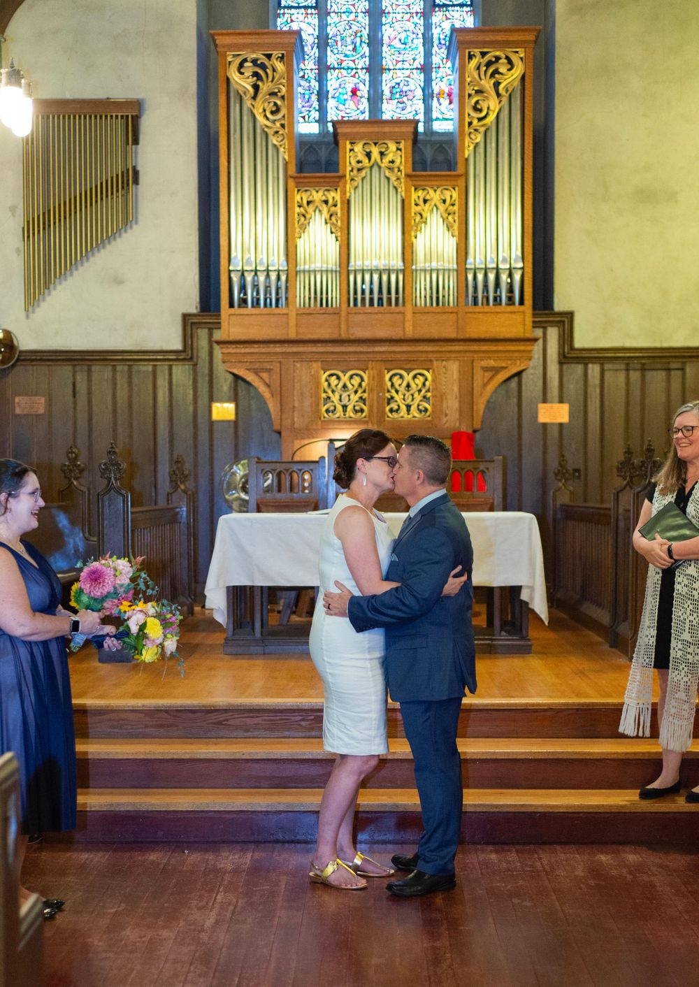 A wedding couple shares a kiss at the altar in a church with an ornate gold organ pipe display behind them.