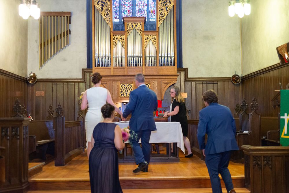 A group gathers around a small altar table with an organ in the background during a church ceremony.