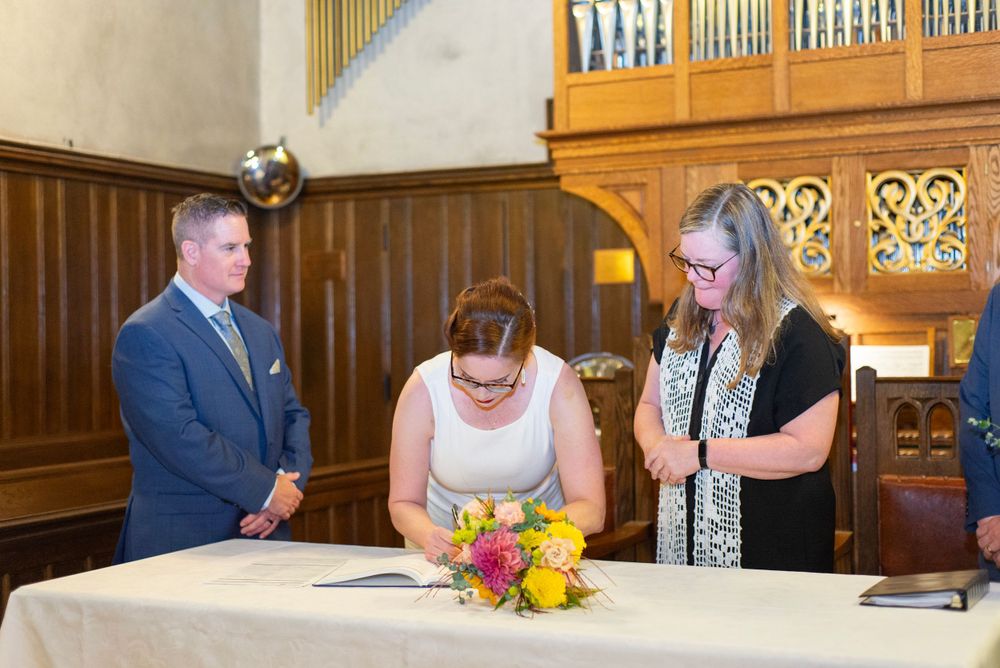 Religious ceremony participants gather at an altar with bright flowers while signing documents inside a church with an organ.