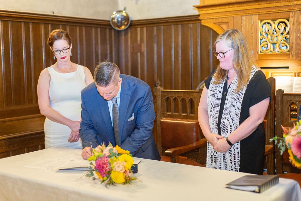 People gather around a wooden table with a colorful floral arrangement during a ceremony in an elegant wood-paneled room.