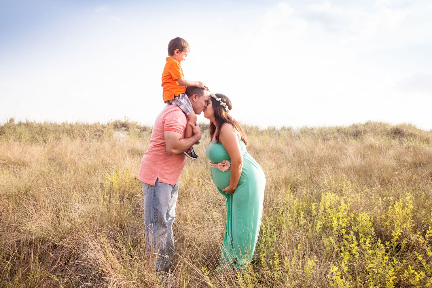 Pregnant woman in a green dress kisses her partner, who carries a young child on his shoulders, in a sunlit grassy field.