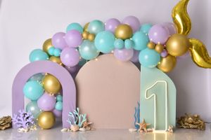 Pastel rainbow balloon garland with gold accents and mermaid-inspired decorative elements against curved panels.