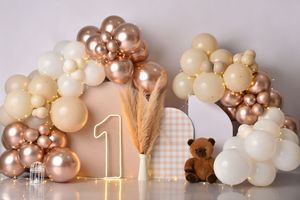 First birthday party backdrop with brown and cream balloon arch, gold number one candle, and teddy bear decorations.