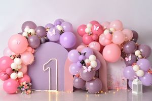 Pink and purple balloon arch with gold accents decorates a first birthday party setup against a gray wall.