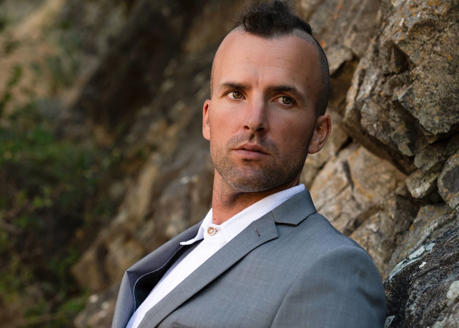 Man in a gray suit posing against a rocky outdoor background.