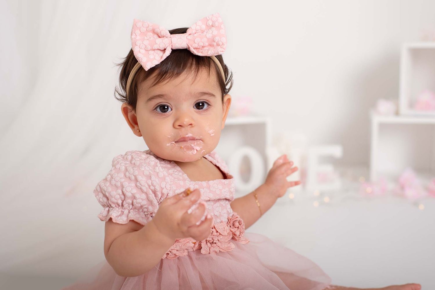 close up baby girl in pink with icing on face for one-year cake smash photo session, celebrating 1st birthday