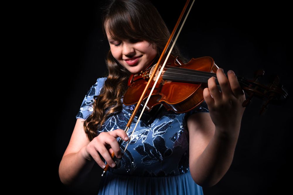 Violinist in blue dress performing against dark background with dramatic lighting.
