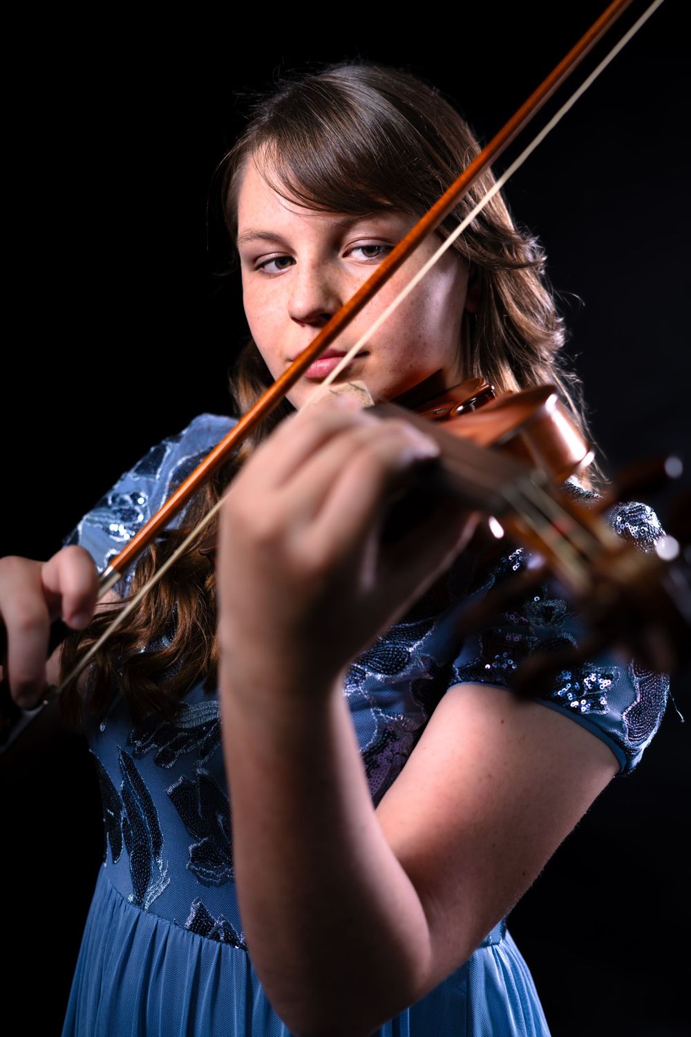 Musician in blue dress plays violin against dark background with dramatic lighting.