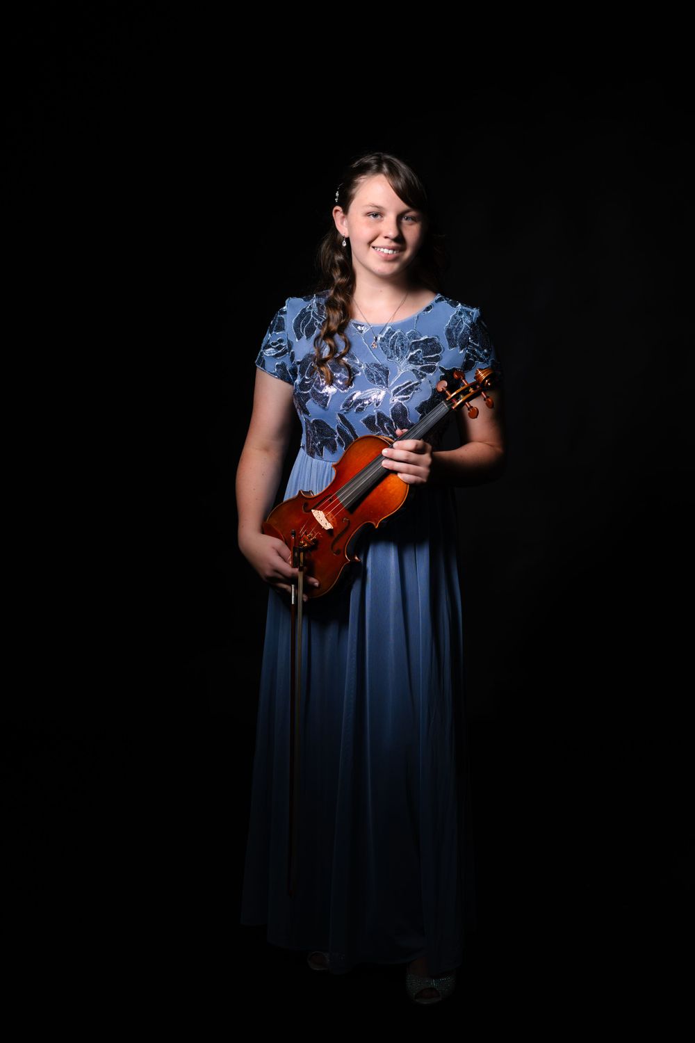 Musician stands holding violin in elegant blue dress against dark studio backdrop.