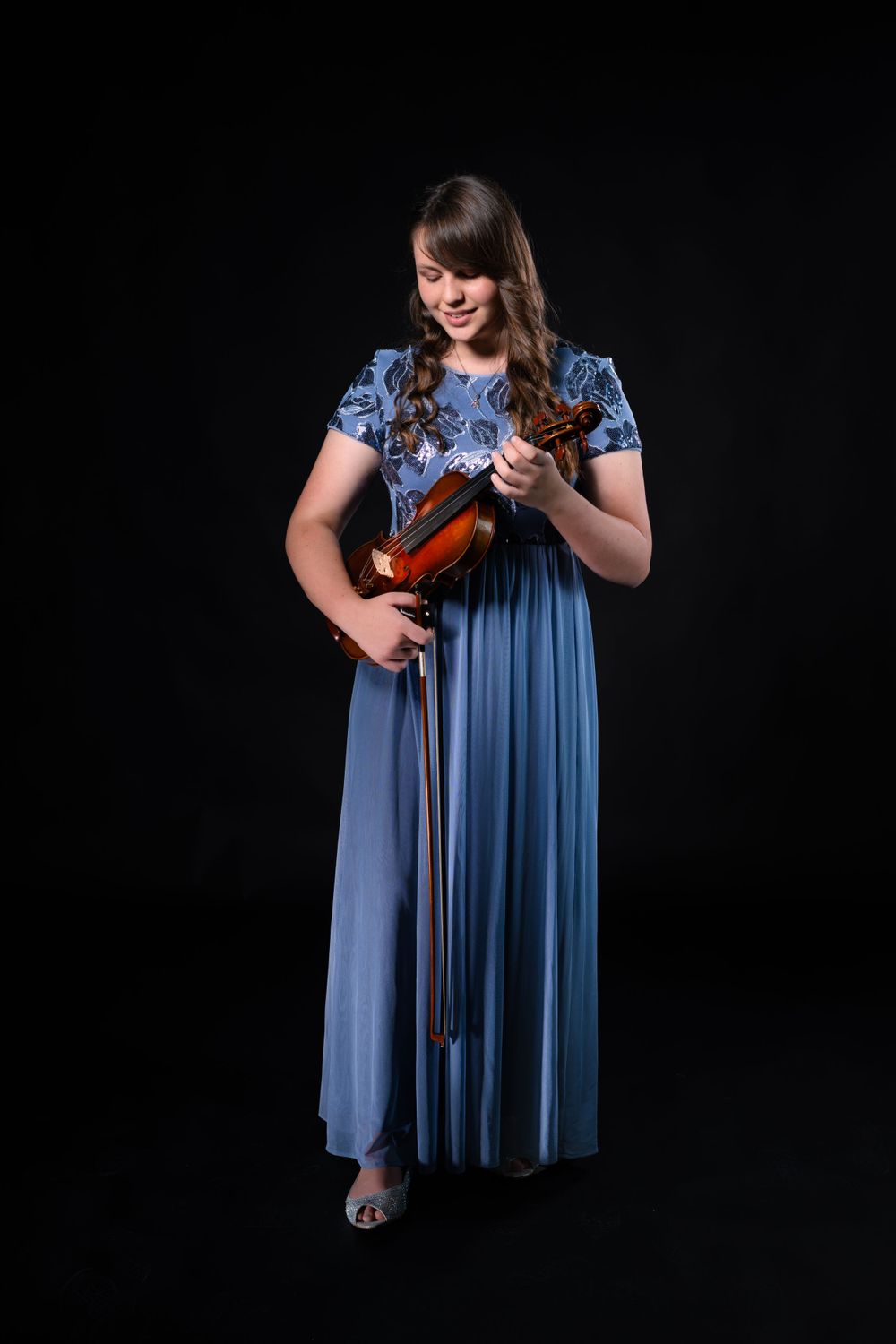 Performer in blue dress adjusts violin strings in dramatic lighting.
