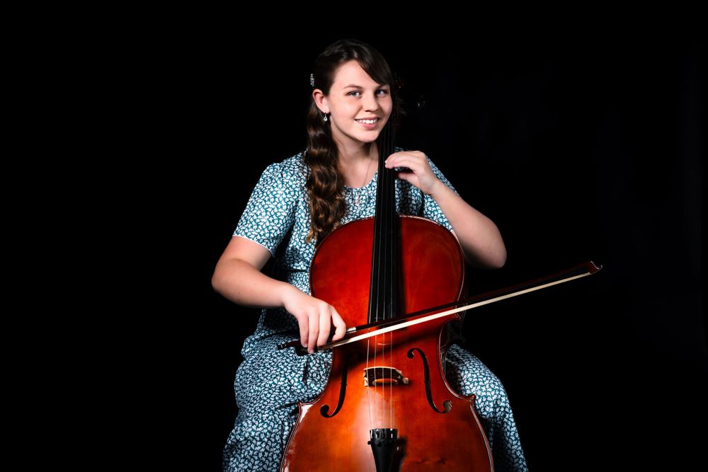 Cellist in patterned dress performs with wooden instrument against black background.