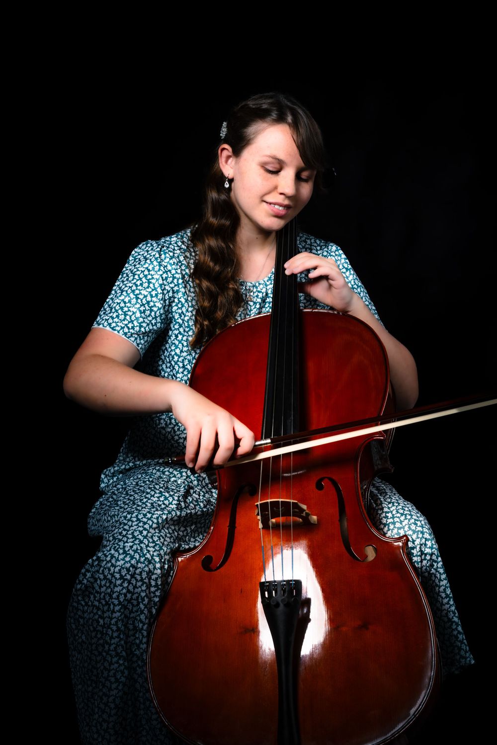 Musician plays cello while wearing spotted blue dress in studio setting.