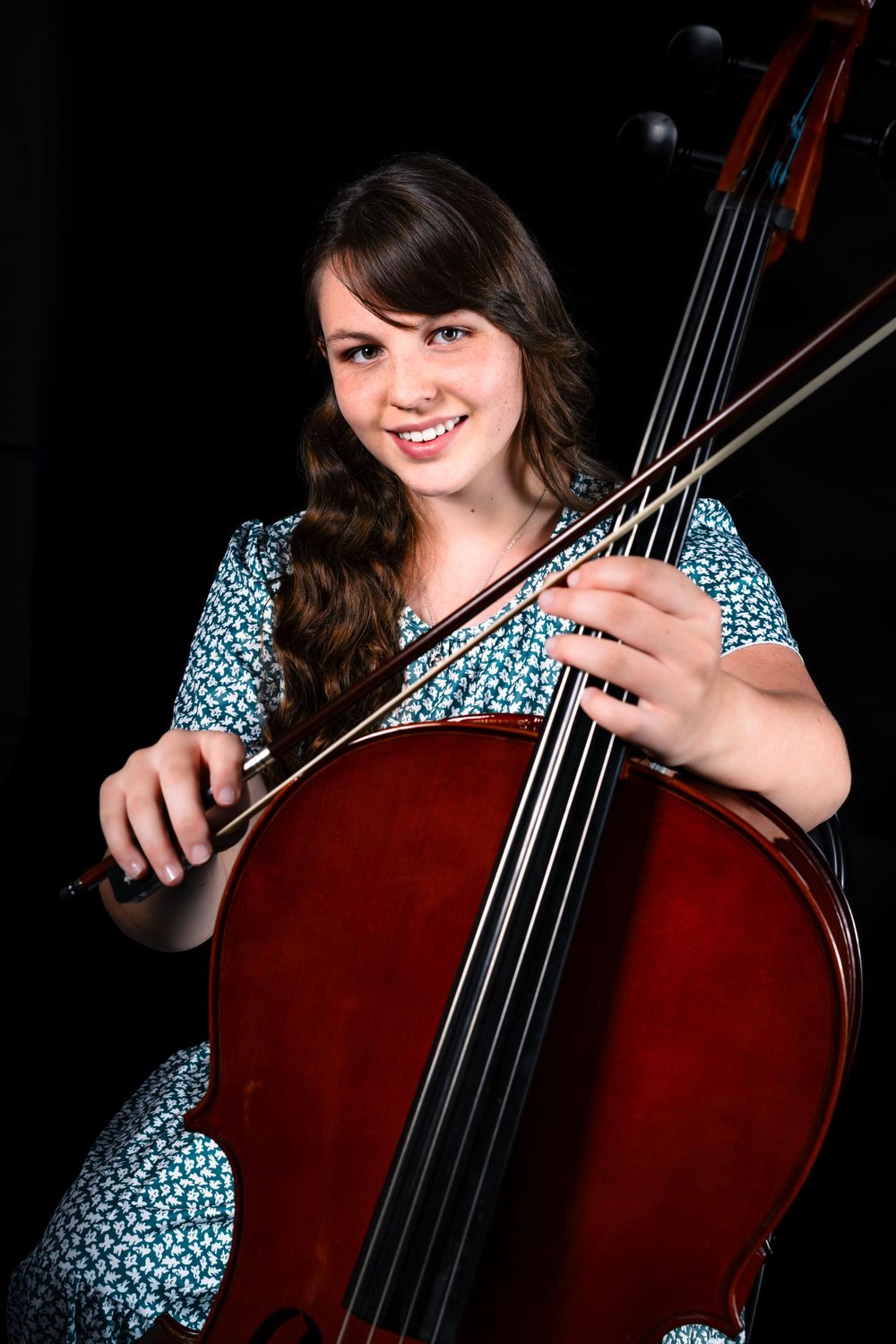 Musician in blue polka dot dress playing a cello against dark background.