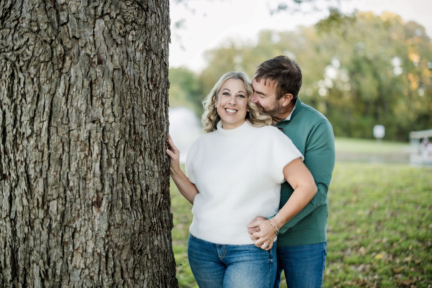 A couple shares an intimate moment while leaning against a large tree trunk during an outdoor photoshoot.