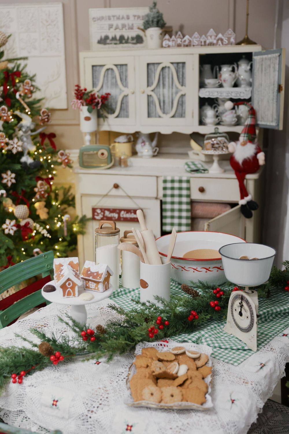 Cozy Christmas kitchen scene with decorated tree, vintage hutch, cookies, and festive greenery on a checkered tablecloth.