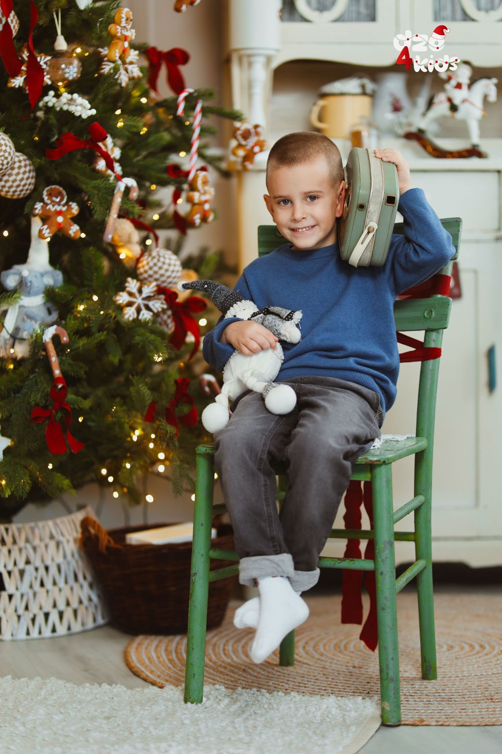 Child sitting on green chair next to decorated Christmas tree holding toys.