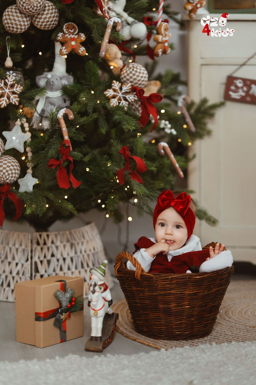 Baby in red outfit sitting in wicker basket by Christmas tree with wrapped presents.