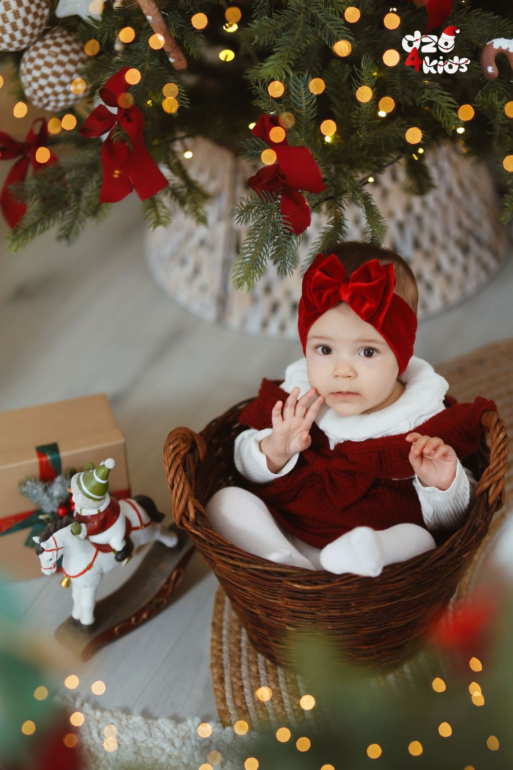 Baby in red holiday outfit sitting in woven basket under lit Christmas tree.