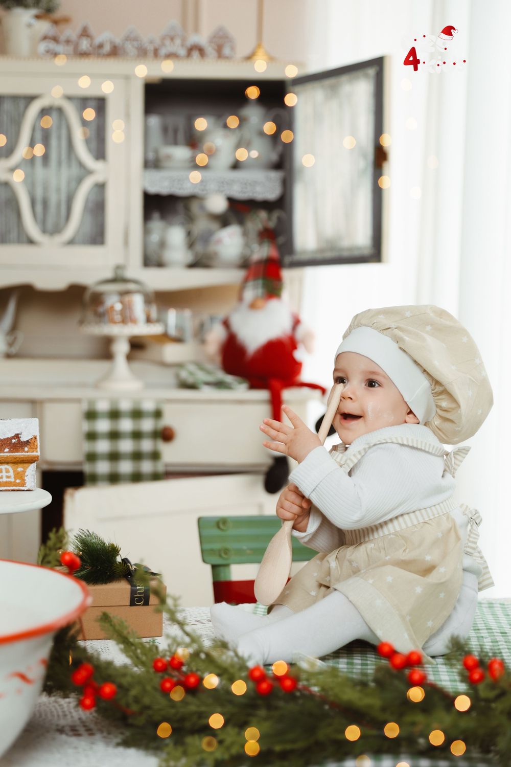Toddler in chef hat exploring Christmas kitchen with decorated hutch and festive lights.
