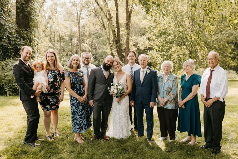 Bride and bearded groom pose with extended family on green lawn beneath shady trees.