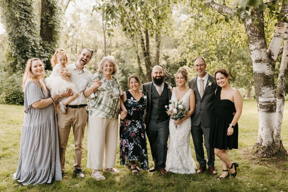 Bride and bearded groom smile with family group outdoors near birch trees in summer.