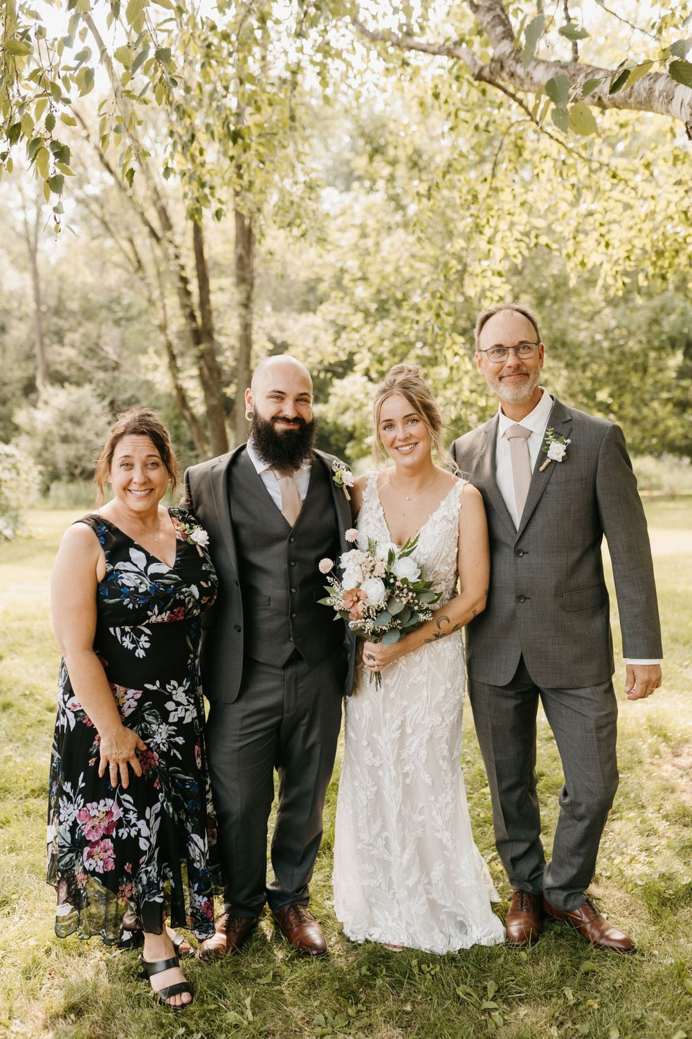 Bride and bearded groom pose with parents outdoors beneath leafy trees on wedding day.