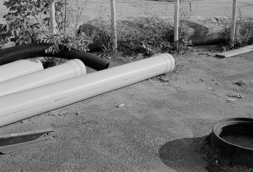 Black and white photograph of white PVC pipes laying on dirt ground next to a dark circular container.