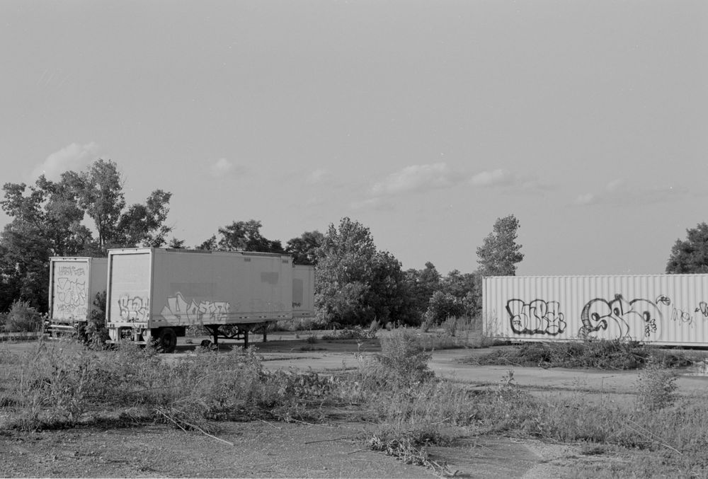 Black and white photograph shows abandoned trailers and containers with graffiti in an overgrown field.