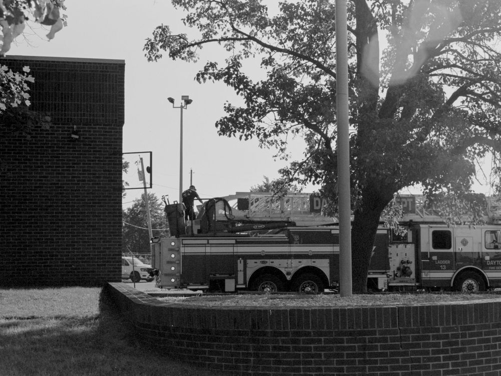A grainy black and white panoramic streetscape showing scattered trees and brick buildings in an urban setting.