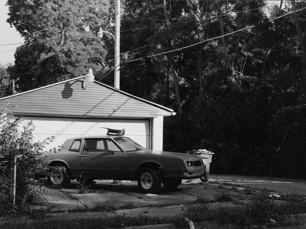 Vintage car and boat trailer parked outside a residential garage surrounded by trees.