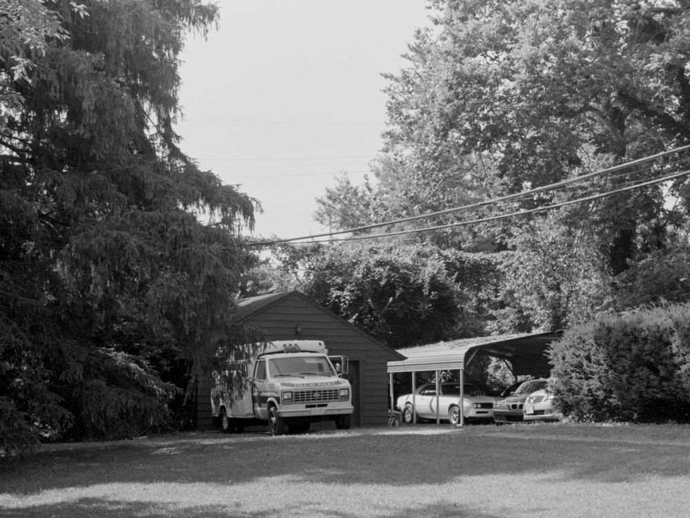 A black and white scenic view of parked vehicles and trees near buildings in a rural or suburban setting.