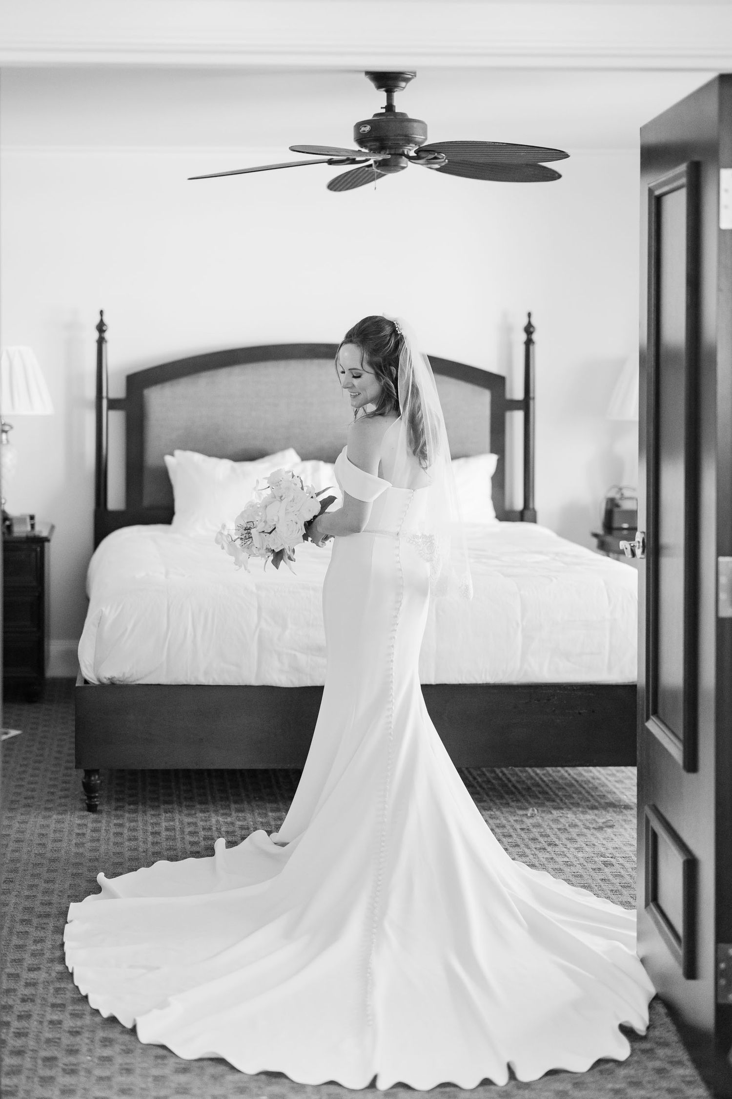 A black and white photo of an elegant wedding dress with a flowing train in a bedroom setting with a ceiling fan.