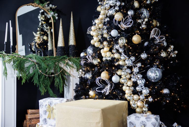 Elegant Christmas tree with gold and white ornaments next to wrapped presents and holiday mantel decor.