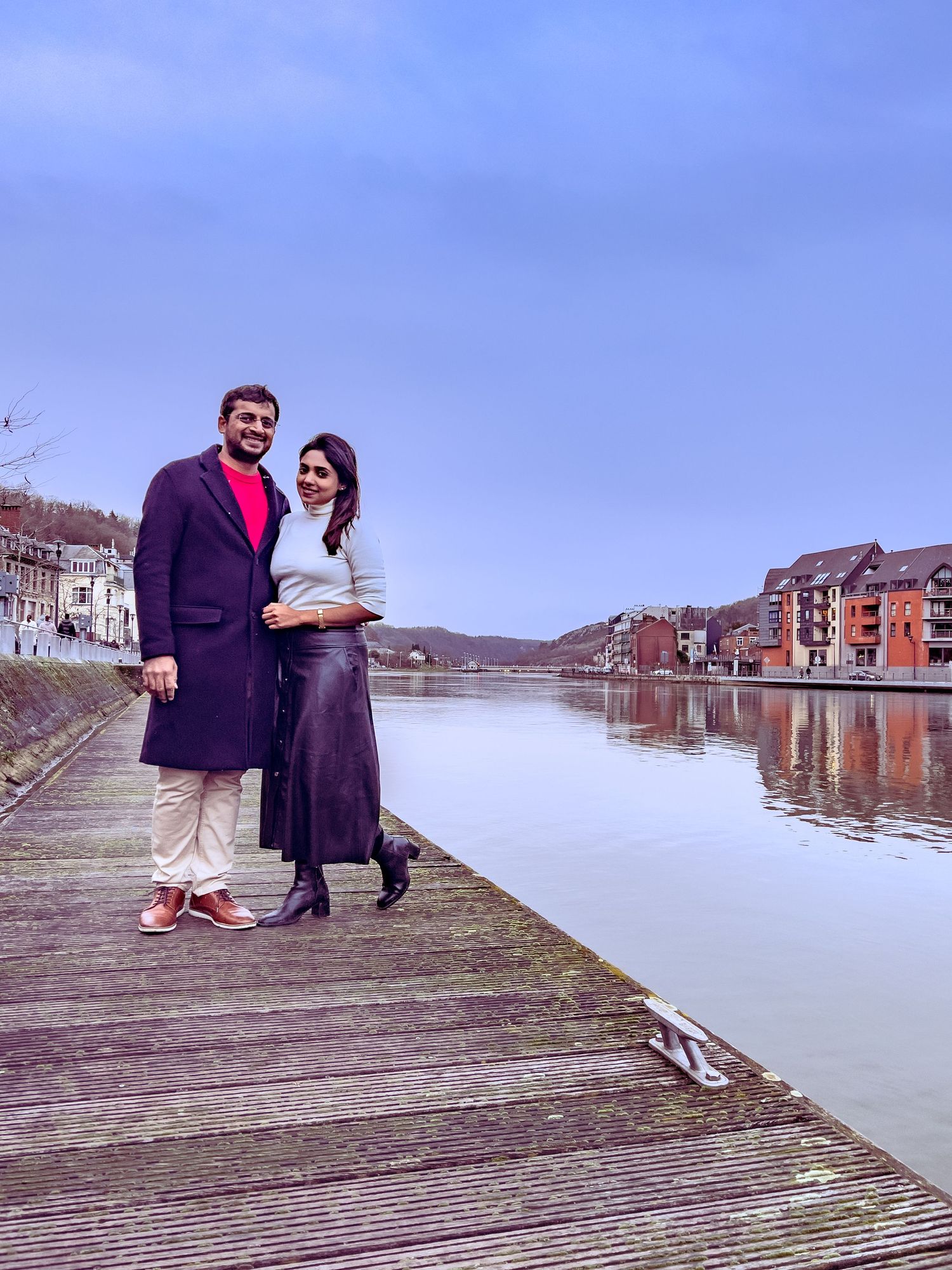 A couple stands together on a wooden boardwalk along a scenic waterfront with historic buildings reflected in calm waters.