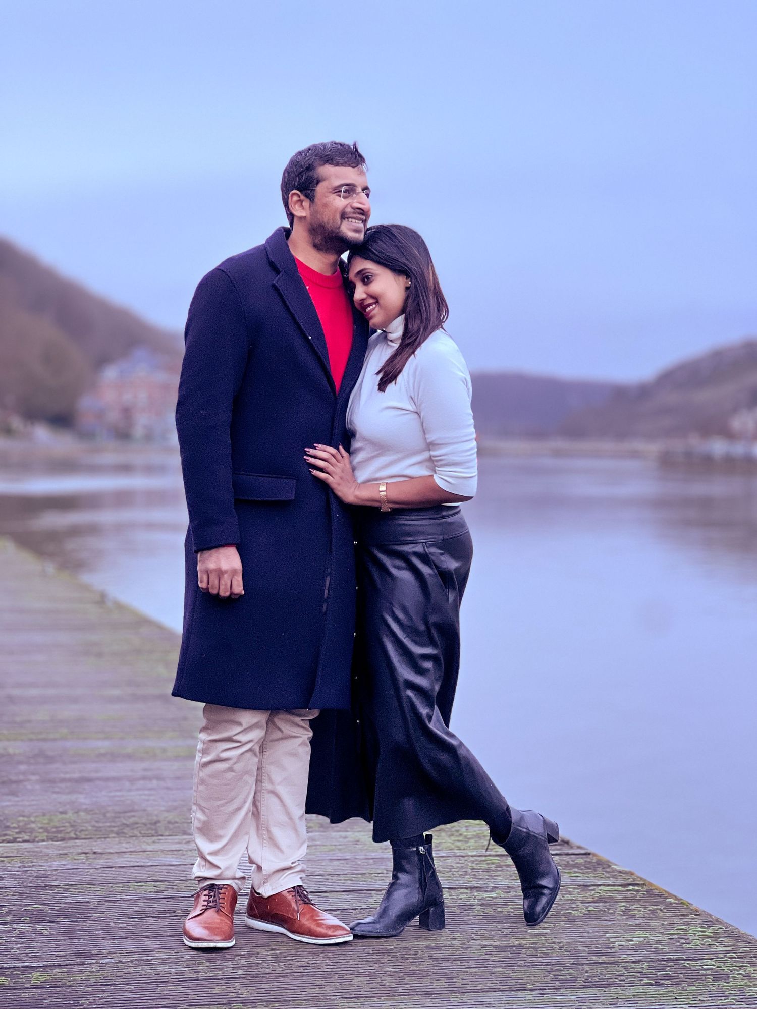 A couple shares an intimate moment while standing on a waterfront path with mountains in the background.