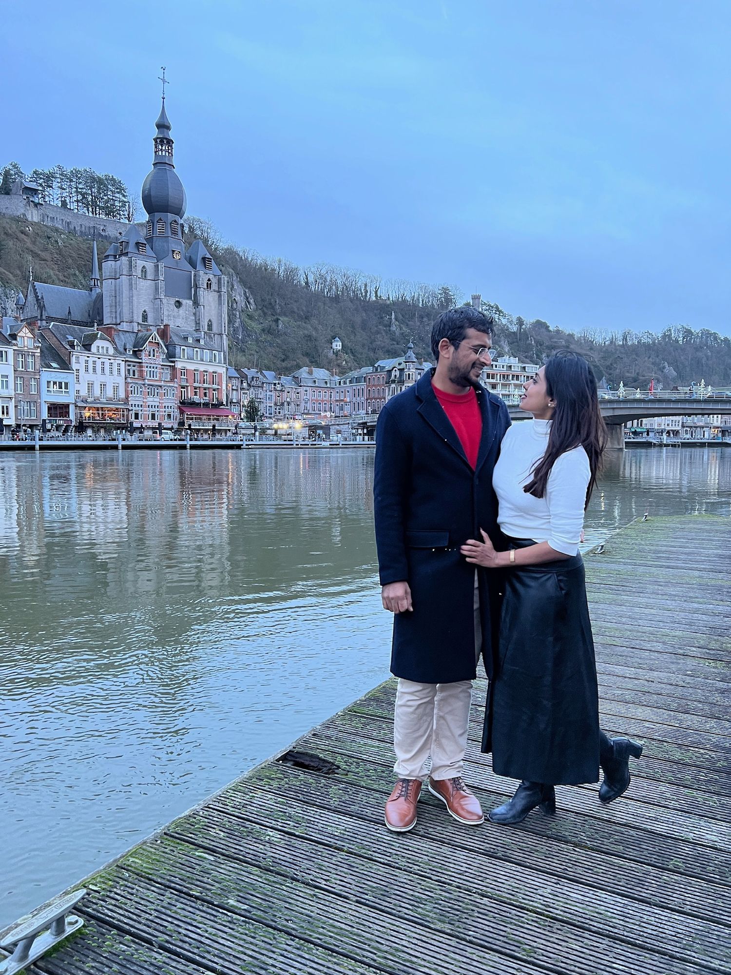 A couple shares a romantic moment on a wooden pier overlooking Dinant, Belgium with its iconic church and cliffside in background.