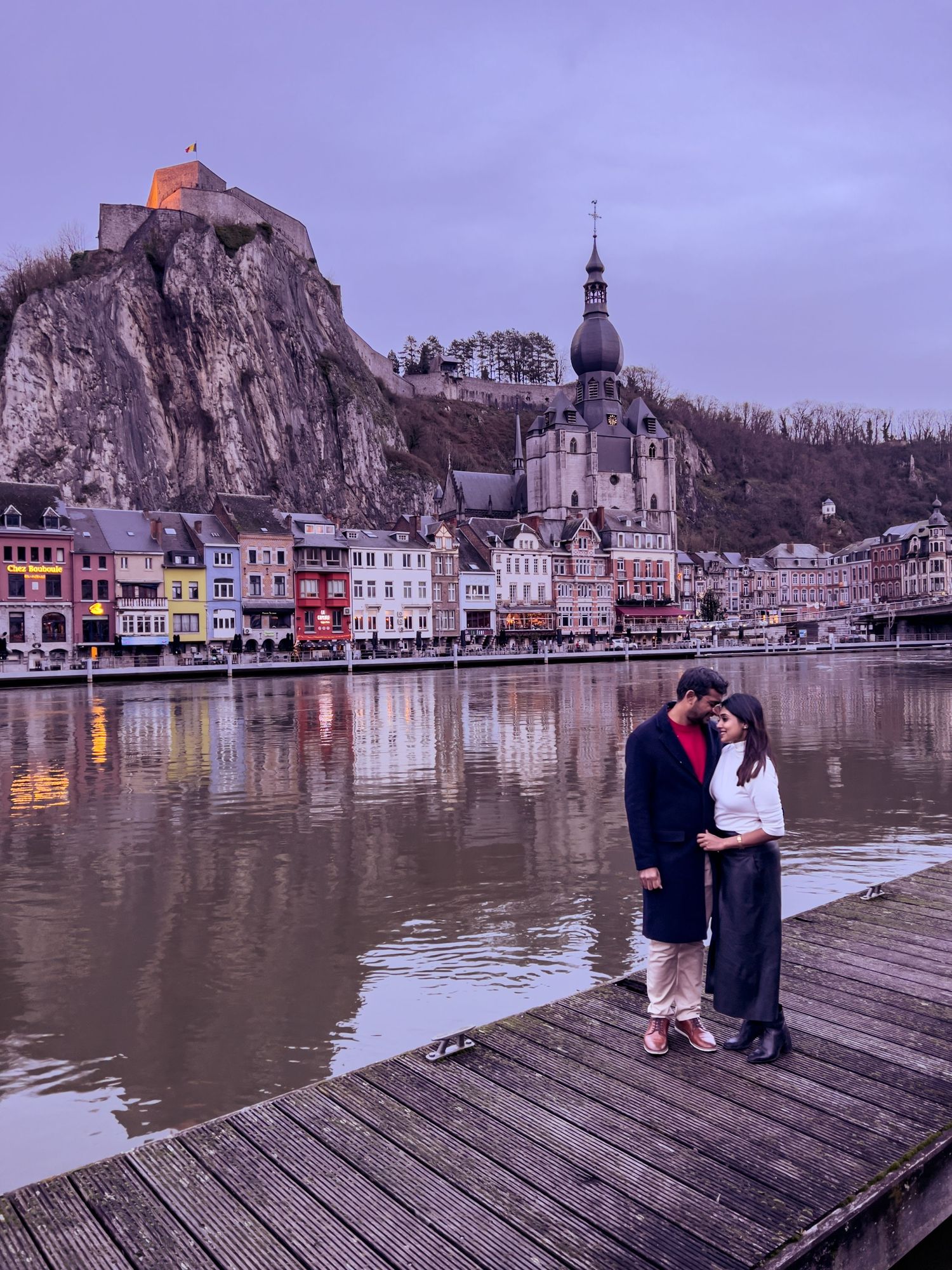 Romantic couple stands on wooden pier overlooking Dinant, Belgium with its iconic citadel and colorful riverside buildings at dusk.