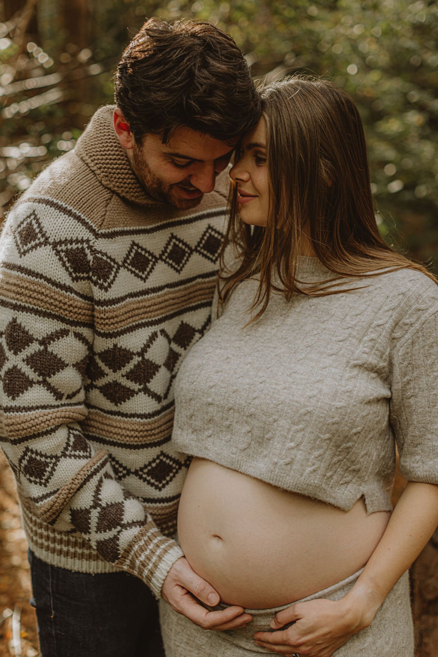 Expectant couple in cozy sweaters share an intimate moment in a forest setting.