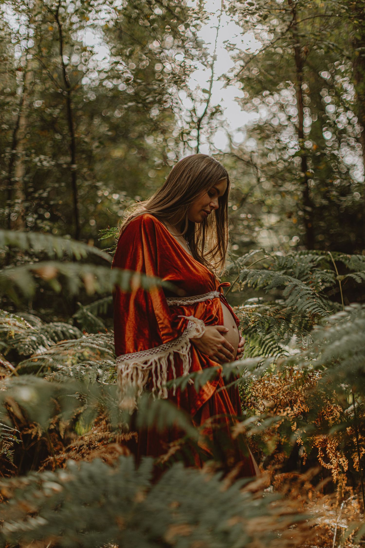 Pregnant woman in red dress standing among ferns in a mystical forest setting.
